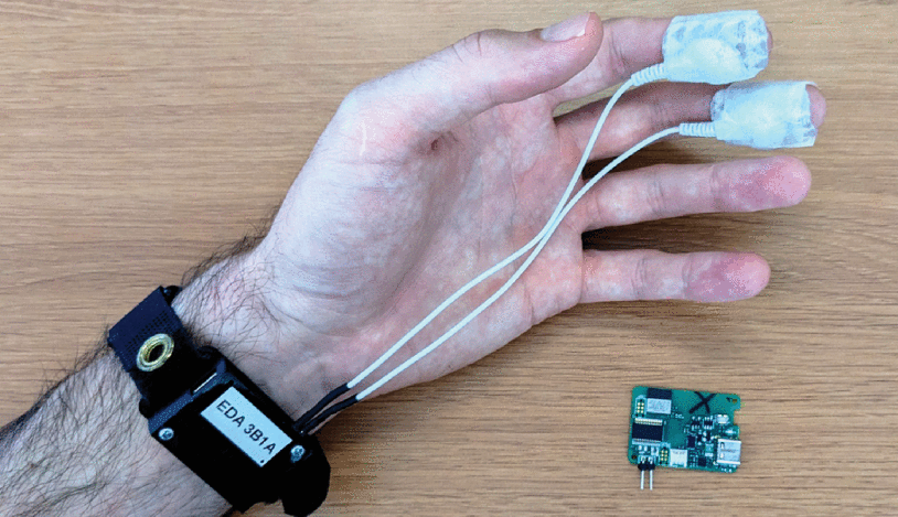 A research setup measuring electrodermal activity (EDA) with electrodes on a subject's hand, connected to an impedance analyzer displaying real-time multi-frequency impedance data